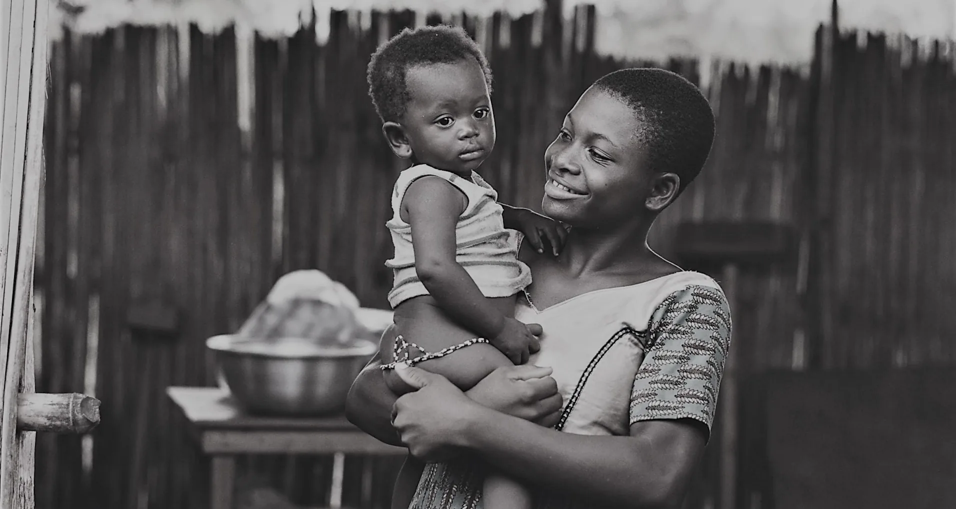 A young mother smiling as she holds her baby on her hip outside a reed-walled home