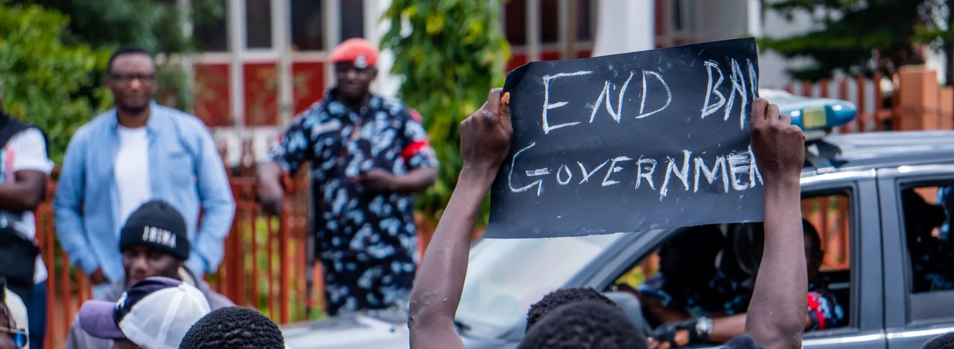 A demonstrator at a street protest in Nigeria holding up a hand-lettered chalkboard placard reading END BAD GOVERNMENT, with a police officer and other protesters behind him