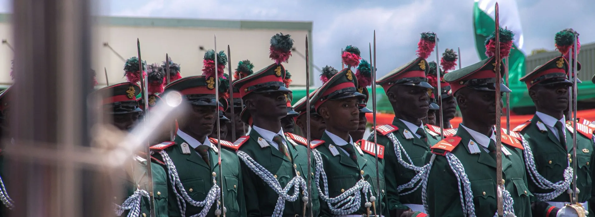 Nigerian officer cadets in green ceremonial dress standing at attention on parade, the national flag behind them