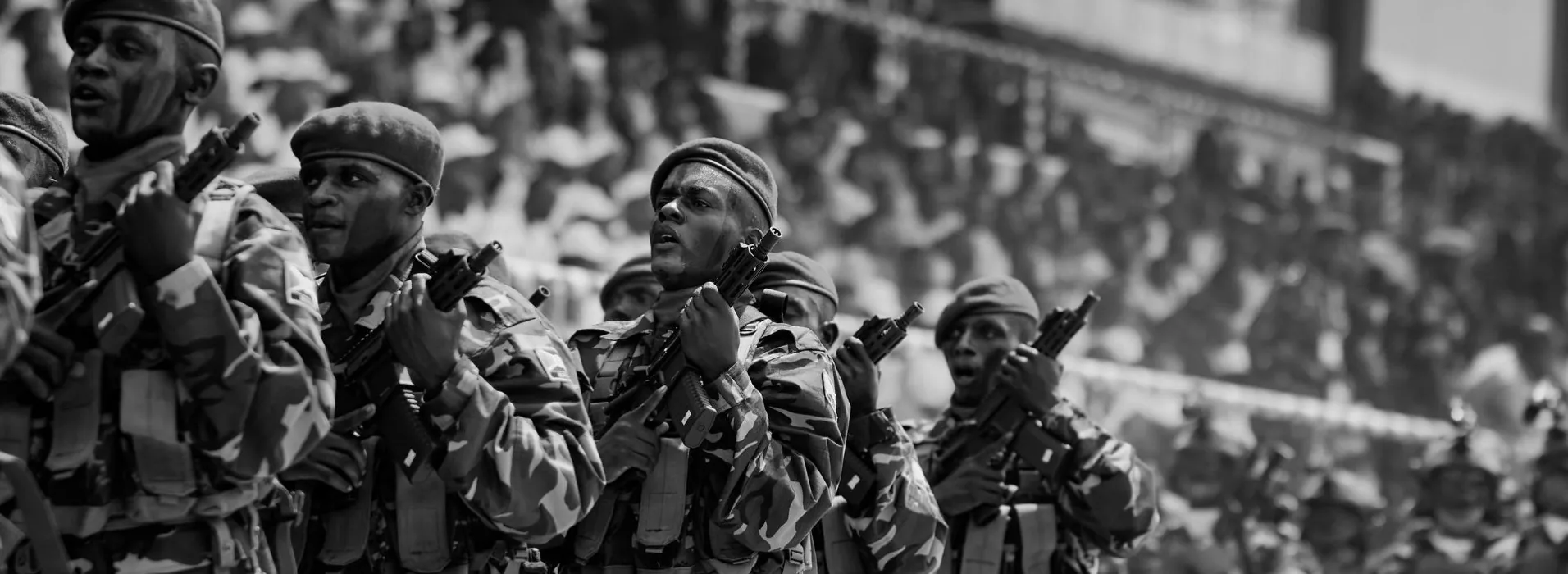 Soldiers in camouflage fatigues and berets marching in column with rifles shouldered, a packed stadium crowd behind them