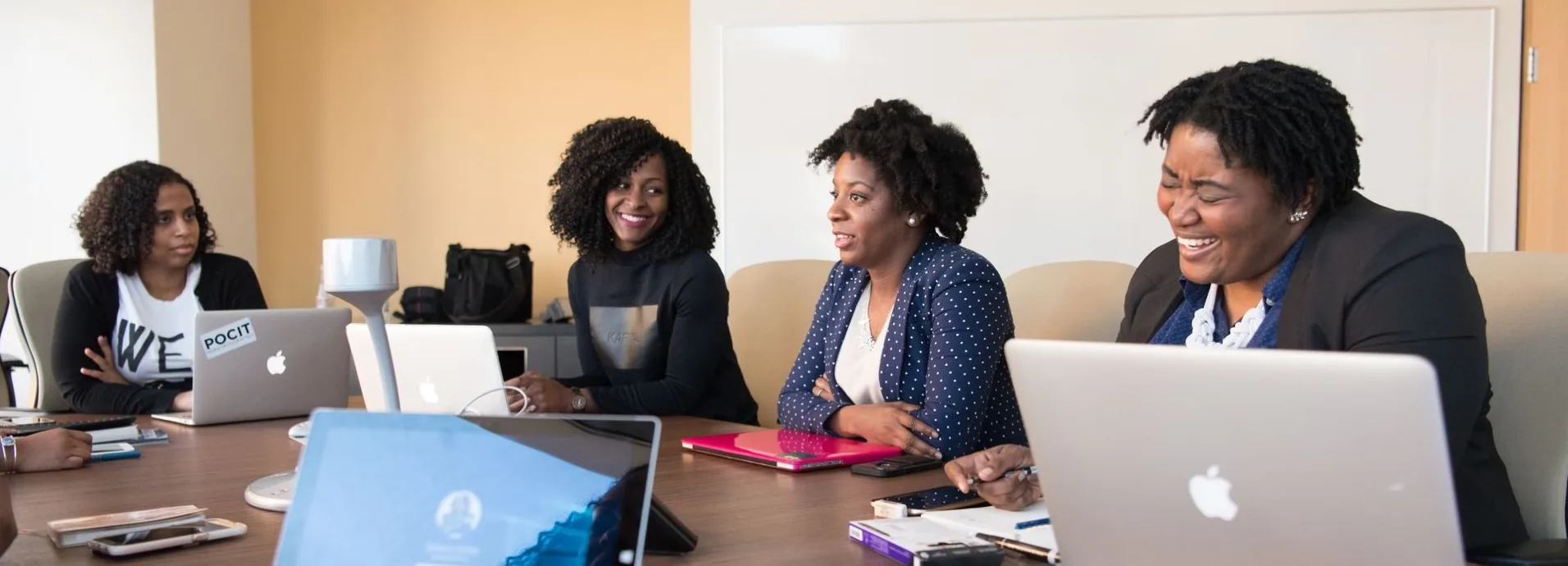Four women meeting around a table with laptops, talking and laughing together