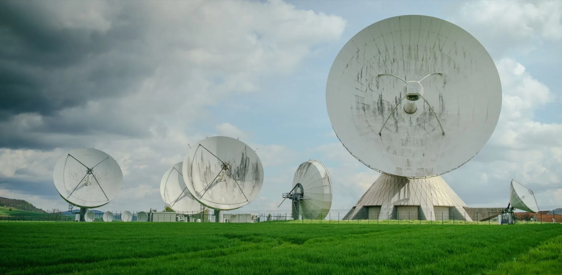 A field of satellite ground-station dishes standing in green grassland under a broken sky