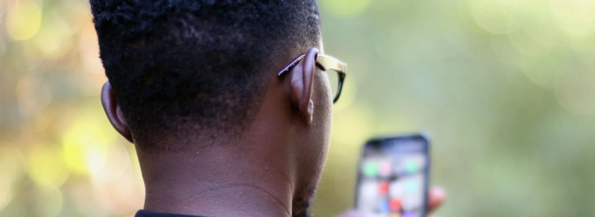 A man in Kenya holding a smartphone, seen from behind against sunlit foliage