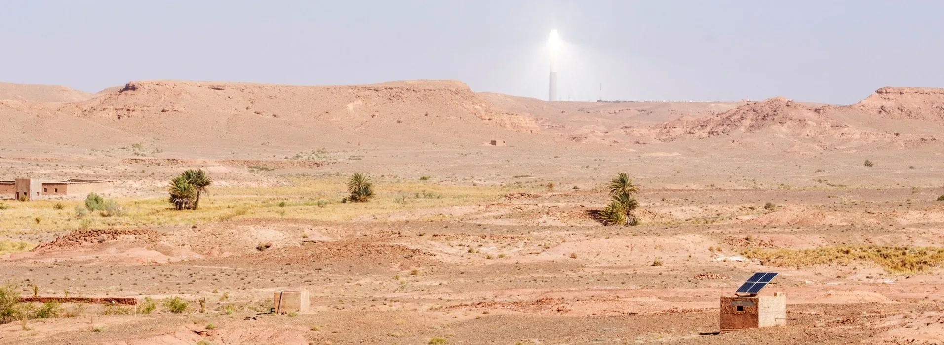 A solar power tower blazing on the horizon of the Moroccan desert, with a small solar panel powering a lone building in the foreground
