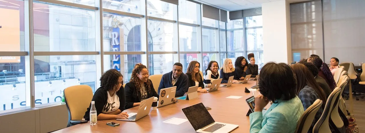 A long meeting table in a glass-walled office, a dozen people seated around it with laptops open, mid-briefing