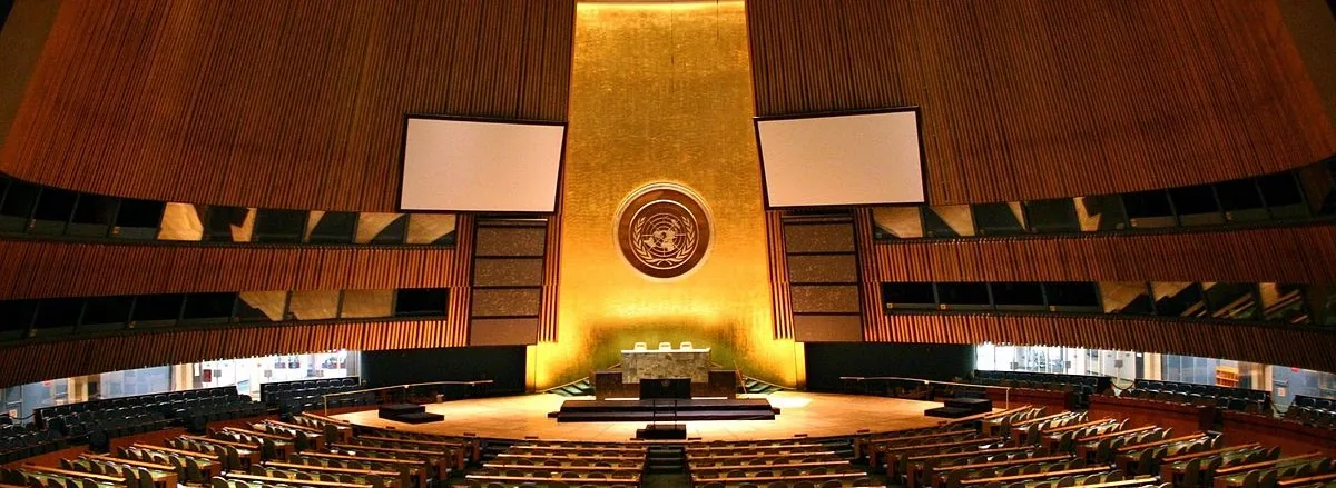 The United Nations General Assembly hall, empty — curved tiers of delegate desks facing the rostrum beneath the UN emblem