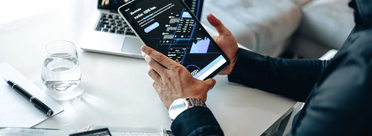 Hands holding a tablet showing a financial tracking dashboard, a laptop with candlestick charts open behind it