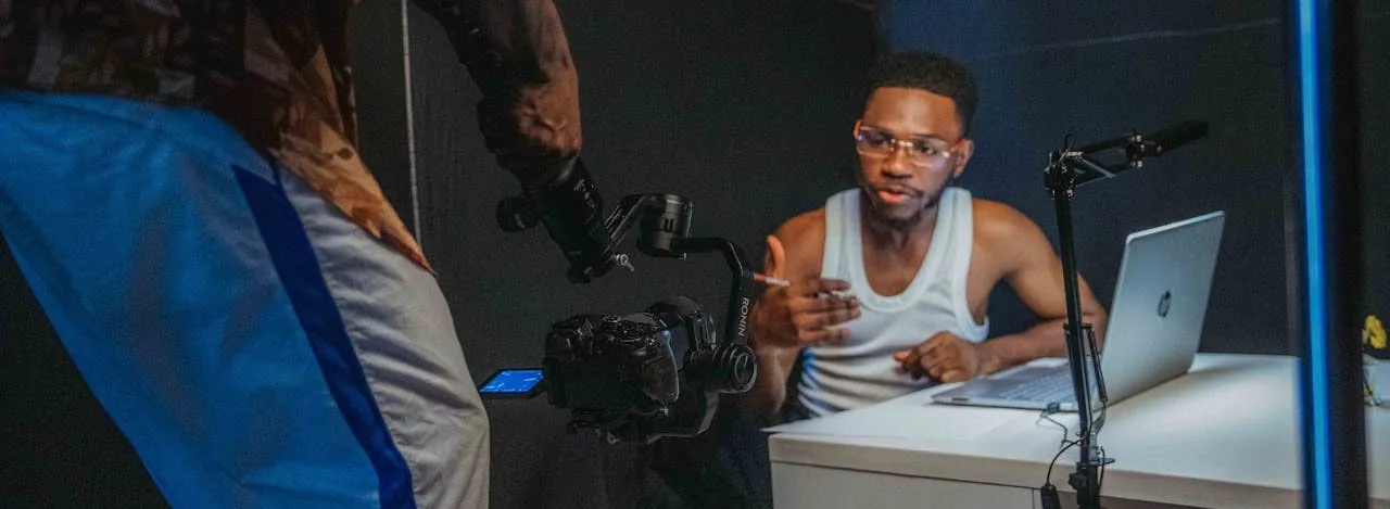 A young man at a desk in a home studio talking to camera, a gimbal-mounted camera and boom microphone rigged in front of him