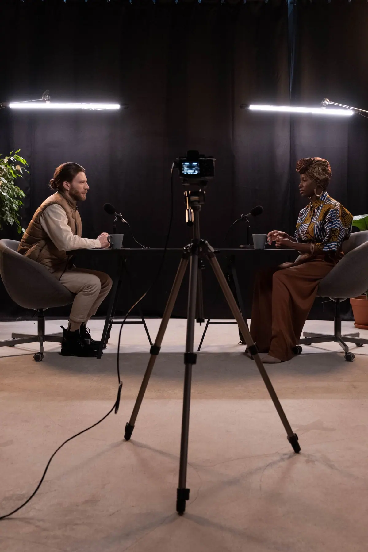 Two hosts recording a filmed interview across a table in a studio