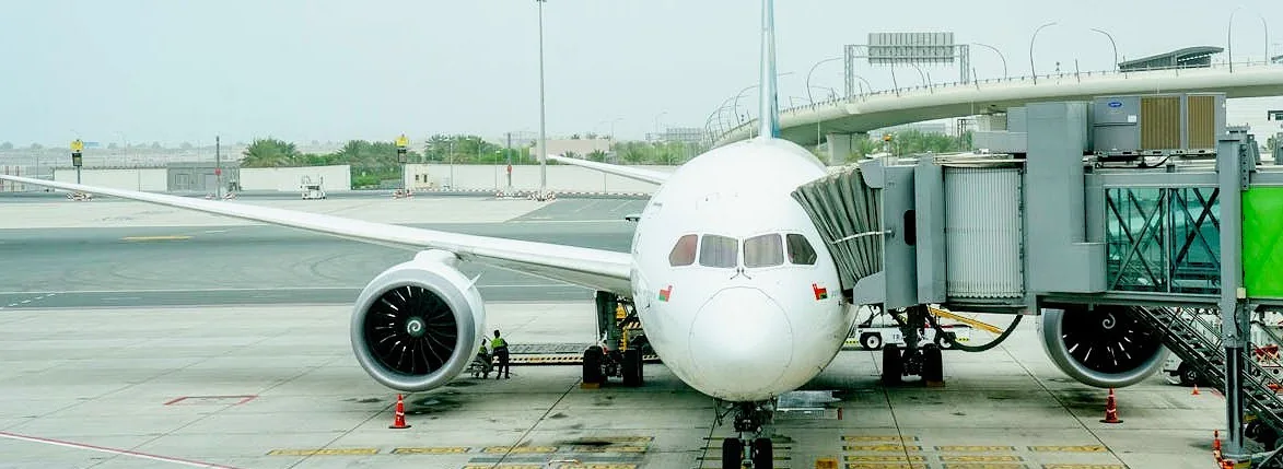 A wide-body airliner parked at a jet bridge on an airport apron, ground crew working beneath the wing