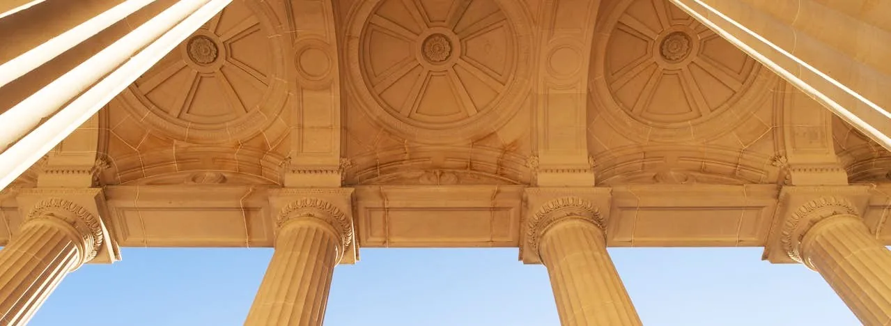 Looking up at the coffered stone portico of a classical institutional building, its columns against a clear sky