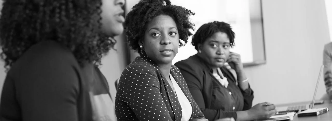Three women seated together in a meeting, listening as a colleague speaks off-camera