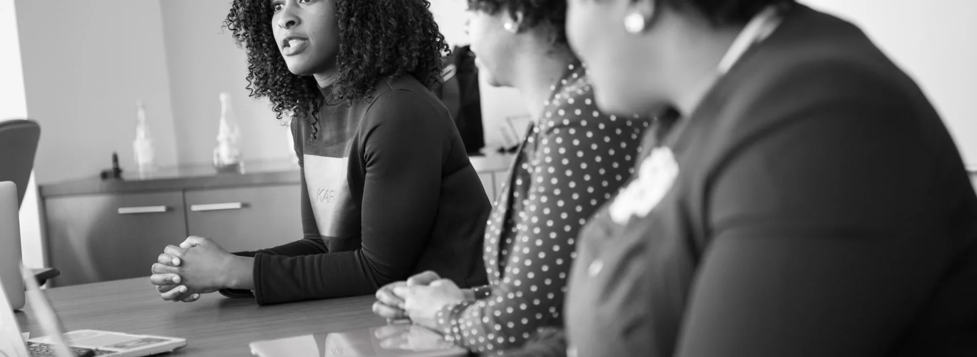 Three women seated around a meeting table mid-discussion, one speaking while the others listen